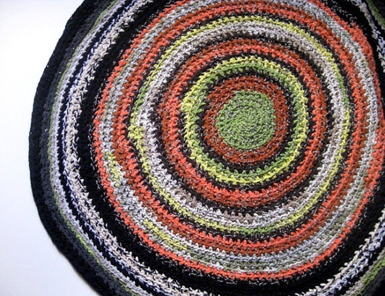 Op de afbeelding: Een ronde, handgemaakte vloerkleed met een spiraalvormig ontwerp. Het vloerkleed heeft concentrische cirkels in verschillende kleuren, waaronder zwart, grijs, wit, oranje, groen en bruin. Het vloerkleed is gemaakt van garen en heeft een getextureerd oppervlak.