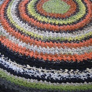 Op de afbeelding: Een handgemaakt, cirkelvormig vloerkleed met een spiraalvormig ontwerp. Het vloerkleed heeft concentrische ringen in verschillende kleuren, waaronder oranje, groen, zwart, wit en grijs. Het vloerkleed is gemaakt van geweven materiaal.
