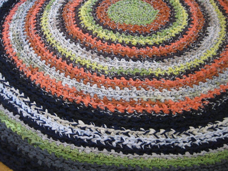Op de afbeelding: Een handgemaakt, cirkelvormig vloerkleed met een spiraalvormig ontwerp. Het vloerkleed heeft concentrische ringen in verschillende kleuren, waaronder oranje, groen, zwart, wit en grijs. Het vloerkleed is gemaakt van geweven materiaal.
