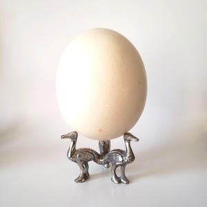 May include: A large, off-white ostrich egg displayed on a silver-colored metal stand. The stand features two stylized bird figures supporting the egg. The egg has a smooth, textured surface. The background is plain white.