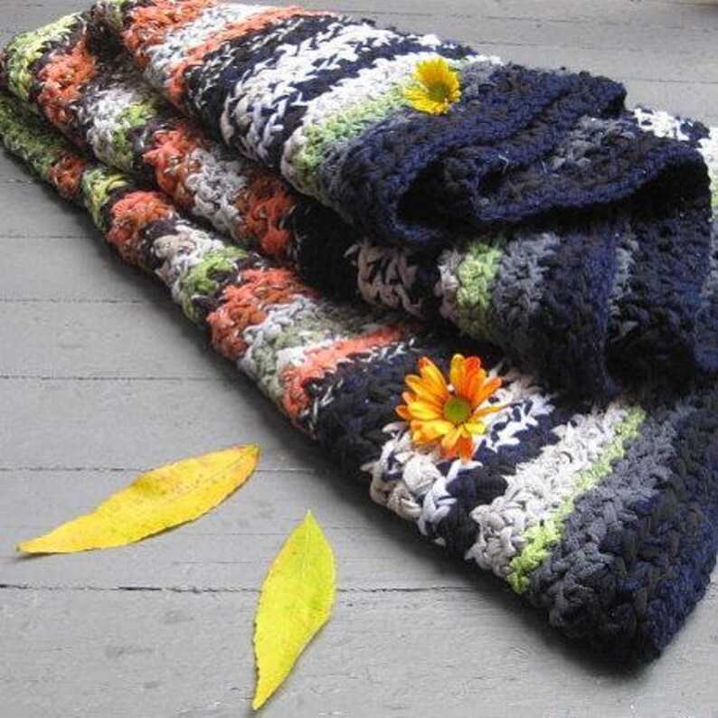 Op de afbeelding: Een gevouwen, handgemaakte gehaakte deken met een gestreept patroon in tinten marineblauw, wit, oranje, groen en grijs. Twee oranje en gele bloemen en twee gele bladeren liggen op de deken.
