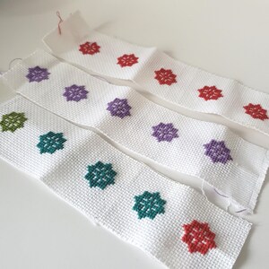 May include: White fabric strips with cross-stitched floral patterns in red, purple, and green. The floral designs are evenly spaced along the strips, creating a repeating pattern. The fabric has a textured appearance.