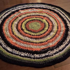 Op de afbeelding: Een ronde, handgemaakte vloerkleed met een spiraalvormig ontwerp. Het vloerkleed heeft concentrische cirkels in verschillende kleuren, waaronder zwart, wit, oranje, groen en geel. Het vloerkleed is gemaakt van een zacht, getextureerd materiaal en is geschikt voor binnenshuis.
