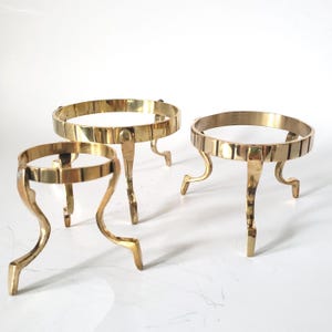 May include: Three polished brass stands, each featuring a circular ring top supported by three curved legs. The stands vary slightly in size, with a smooth, reflective metallic finish and a simple, functional design for holding spherical objects.