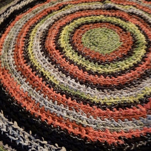 Op de afbeelding: Een ronde, handgemaakte vloerkleed met een spiraalvormig ontwerp. Het vloerkleed heeft concentrische cirkels in verschillende kleuren, waaronder zwart, koraal, groen en grijs. Het vloerkleed is gemaakt van een geweven materiaal en de textuur is zichtbaar.