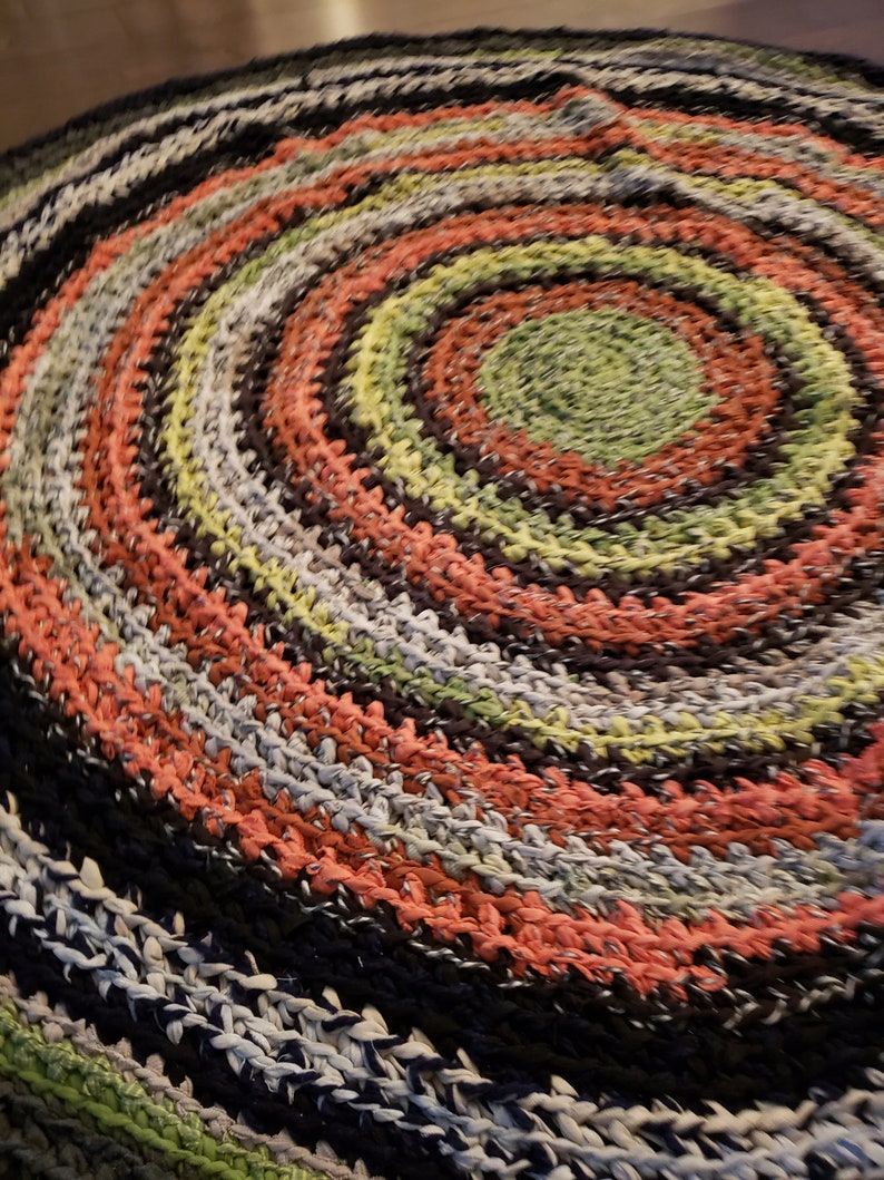 Op de afbeelding: Een ronde, handgemaakte vloerkleed met een spiraalvormig ontwerp. Het vloerkleed heeft concentrische cirkels in verschillende kleuren, waaronder zwart, koraal, groen en grijs. Het vloerkleed is gemaakt van een geweven materiaal en de textuur is zichtbaar.