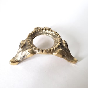May include: A small, antique brass stand with a circular opening in the centre. The stand has three feet and a decorative, textured rim. The brass has a warm, golden hue.