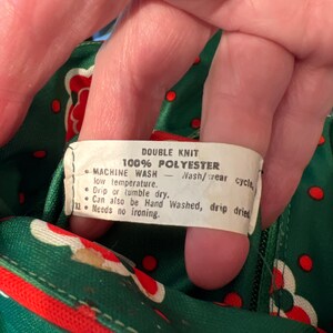 Pu&ograve; includere: Un'etichetta bianca con testo nero su uno sfondo di tessuto verde e rosso. L'etichetta recita "Double Knit 100% Polyester Machine Wash - Nash/wear cycle, low temperature. Drip or tumble dry. Can also be Hand Washed, drip dried. Needs no ironing."