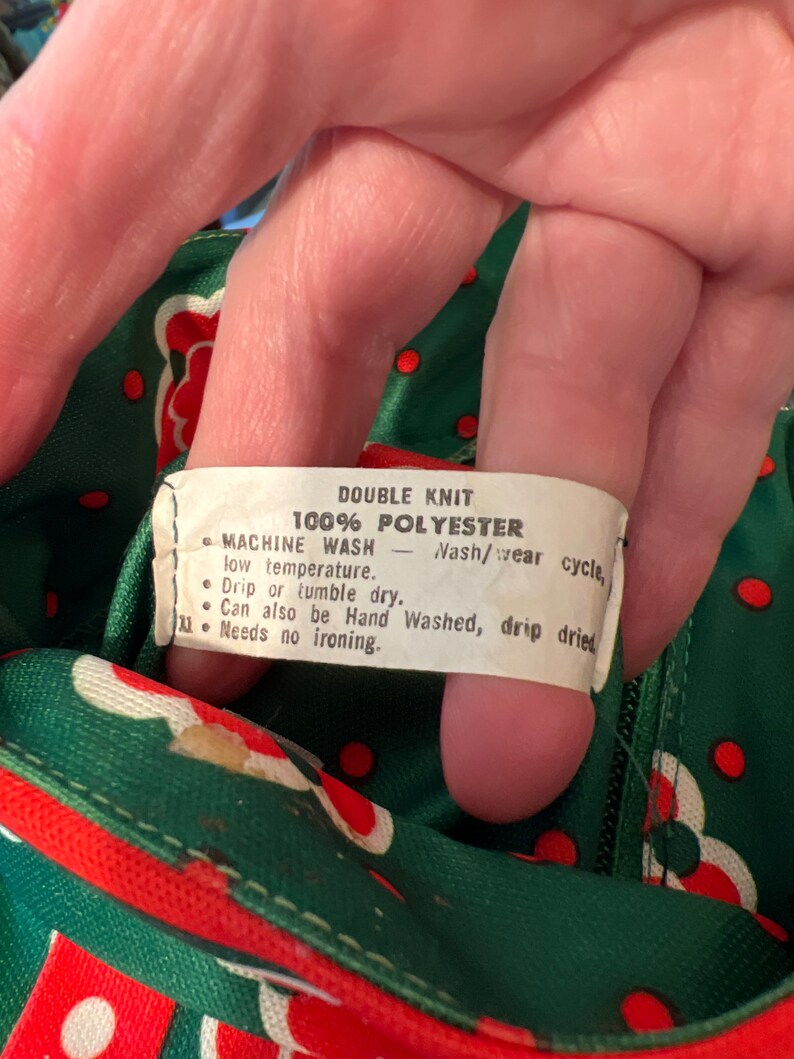 Pu&ograve; includere: Un'etichetta bianca con testo nero su uno sfondo di tessuto verde e rosso. L'etichetta recita "Double Knit 100% Polyester Machine Wash - Nash/wear cycle, low temperature. Drip or tumble dry. Can also be Hand Washed, drip dried. Needs no ironing."