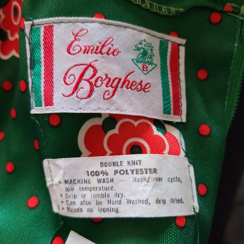 Pu&ograve; includere: Un'etichetta bianca con bordino rosso e verde, con il testo "Emilio Borghese" in lettere rosse corsive. L'etichetta include anche un piccolo logo ricamato di una testa di cavallo. L'etichetta &egrave; attaccata a un tessuto verde con pois rossi e un motivo floreale rosso. L'etichetta include le istruzioni di cura per il tessuto: "Double Knit 100% Polyester Machine Wash - Nash/wear cycle, low temperature. Drip or tumble dry. Can also be Hand Washed, drip dried. Needs no ironing."