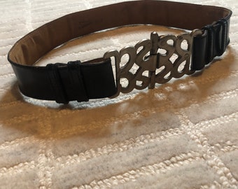 May include: A black leather belt with a silver buckle featuring an ornate design. The buckle is shaped like a stylized word.