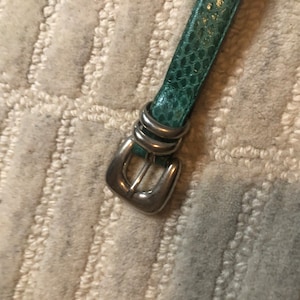 Könnte beinhalten: Ein türkisfarbener Ledergürtel mit einer silbernen Schnalle. Die Schnalle ist ein schlichtes, rechteckiges Design mit einem einzigen Stift.