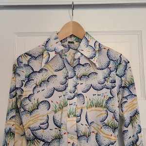 Puede incluir: Una camisa blanca de manga larga con un patrón de nubes azules y blancas. La camisa tiene un cuello puntiagudo y un cierre de botones en la parte delantera.