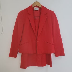 Könnte beinhalten: Ein roter Blazer und Rock-Anzug. Der Blazer hat einen einzigen Knopfverschluss und zwei Taschen. Der Rock ist knielang.