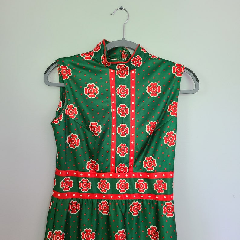 Pu&ograve; includere: Un abito verde senza maniche con un motivo floreale rosso e bianco. L'abito ha un collo alto e un corpetto aderente. La gonna &egrave; ampia e fluida.