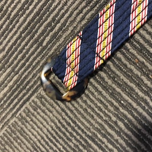 May include: A close-up of a navy blue and red striped belt with a tortoiseshell buckle. The belt is made of a woven fabric and has a subtle sheen.