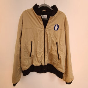 May include: A beige bomber jacket with a black zipper and black ribbed cuffs. The jacket has a blue and white patch with a bear on the left side and a green and white "IZOD LACOSTE" label on the right side.