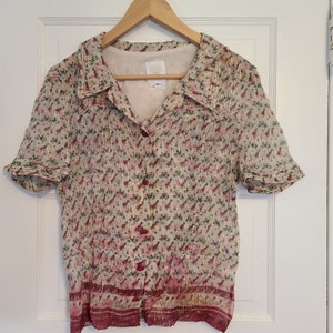 May include: A short-sleeved, button-up shirt with a collared neckline. The shirt is made of a light-weight, sheer fabric with a floral print and gold accents. The shirt has a red and gold trim around the bottom.