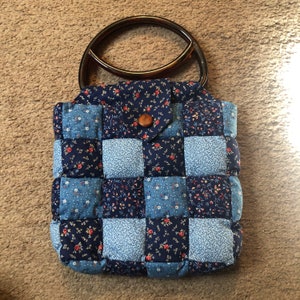 May include: A blue and white quilted handbag with a brown wooden handle. The bag is made of patchwork fabric with a floral pattern.