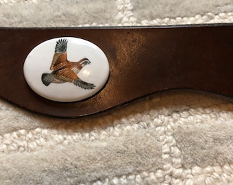 May include: A brown leather belt with a white oval buckle featuring a painted image of a brown and white quail in flight.
