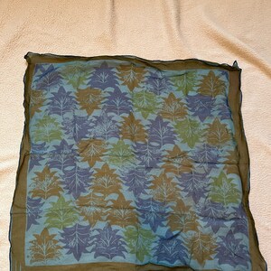 May include: A blue, green, and brown patterned silk scarf with a dark brown border and the word "Una" in the bottom left corner.