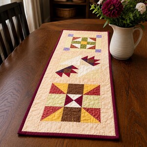 Peut inclure: Un chemin de table matelassé rectangulaire avec un fond crème et une bordure rouge foncé. Le chemin présente trois blocs en patchwork distincts dans des tons de marron, jaune, vert, rouge et blanc, avec des motifs géométriques et de pattes d'ours.