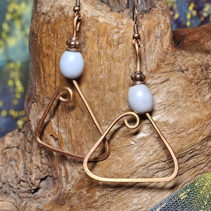May include: Copper wire earrings with a triangular design. Each earring features a pale blue bead and a hook closure. The earrings are displayed on a piece of wood with a colorful fabric background.