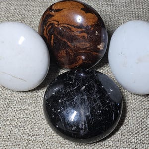 May include: Four polished stones: two white, one brown with dark veining, and one black with white streaks. The stones are smooth and egg-shaped, arranged on a textured, neutral-colored surface. These decorative stones are ideal for home decor.