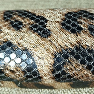 May include: An oval-shaped eyeglasses case with a leopard print design. The case has a tan and brown pattern with black mesh accents and silver sequins. It is designed to protect eyeglasses.