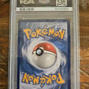 Puede incluir: Una carta de Pok&eacute;mon clasificada, encapsulada en un estuche de pl&aacute;stico transparente. El reverso de la carta presenta un dise&ntilde;o arremolinado azul y blanco con la palabra "Pok&eacute;mon" en naranja y amarillo. El estuche tiene el logotipo de PSA y un c&oacute;digo de barras.