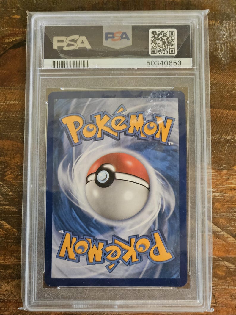 Puede incluir: Una carta de Pok&eacute;mon clasificada, encapsulada en un estuche de pl&aacute;stico transparente. El reverso de la carta presenta un dise&ntilde;o arremolinado azul y blanco con la palabra "Pok&eacute;mon" en naranja y amarillo. El estuche tiene el logotipo de PSA y un c&oacute;digo de barras.