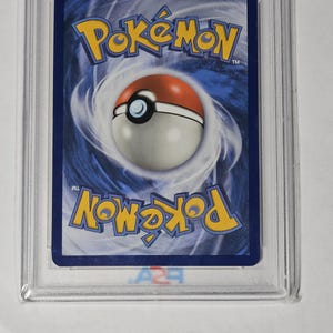 May include: A graded Pok&eacute;mon trading card, encased in a clear plastic holder. The card's back features a blue background with the Pok&eacute;mon logo in yellow and a Pok&eacute; Ball design. The card is in a protective case.
