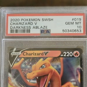 Puede incluir: Una carta Pok&eacute;mon calificada con Charizard V del conjunto Darkness Ablaze. La carta est&aacute; dentro de un soporte protector con el texto "2020 POKEMON SWSH CHARIZARD V DARKNESS ABLAZE" y una calificaci&oacute;n de "GEM MT 10".