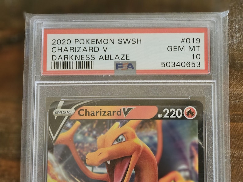 Puede incluir: Una carta Pok&eacute;mon calificada con Charizard V del conjunto Darkness Ablaze. La carta est&aacute; dentro de un soporte protector con el texto "2020 POKEMON SWSH CHARIZARD V DARKNESS ABLAZE" y una calificaci&oacute;n de "GEM MT 10".