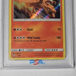 May include: A graded Pok&eacute;mon trading card featuring Charizard. The card has a red and orange background with the image of a dragon-like creature. Text includes "Slash" and "Wild Tackle" with damage values. The card is encased in a protective holder.