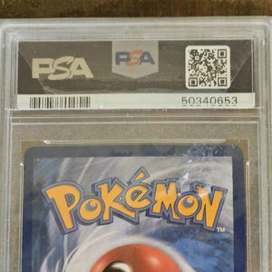 Puede incluir: Una carta coleccionable de Pok&eacute;mon graduada, encapsulada en un soporte de pl&aacute;stico transparente. El soporte presenta el logotipo de PSA, un c&oacute;digo QR y el n&uacute;mero 50340653. El logotipo de Pok&eacute;mon es visible en la carta.