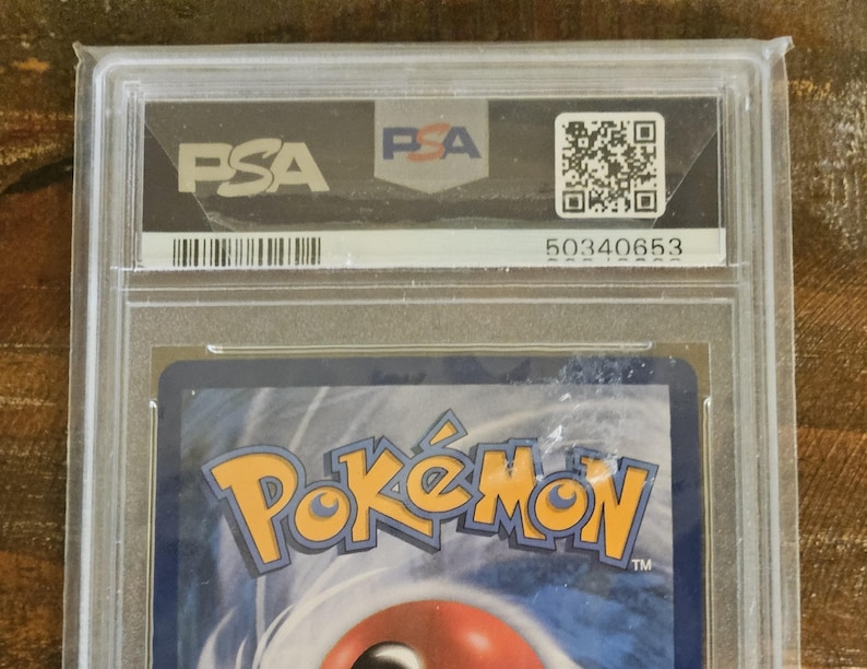 Puede incluir: Una carta coleccionable de Pok&eacute;mon graduada, encapsulada en un soporte de pl&aacute;stico transparente. El soporte presenta el logotipo de PSA, un c&oacute;digo QR y el n&uacute;mero 50340653. El logotipo de Pok&eacute;mon es visible en la carta.