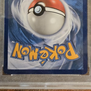 Puede incluir: Una carta coleccionable de Pok&eacute;mon calificada, encerrada en un soporte de pl&aacute;stico transparente. La tarjeta presenta el logotipo de Pok&eacute;mon en amarillo y un dise&ntilde;o de Pok&eacute; Ball. El fondo es un patr&oacute;n azul y blanco arremolinado. La tarjeta est&aacute; sobre una superficie de madera.