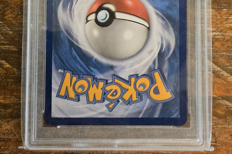 Puede incluir: Una carta coleccionable de Pok&eacute;mon calificada, encerrada en un soporte de pl&aacute;stico transparente. La tarjeta presenta el logotipo de Pok&eacute;mon en amarillo y un dise&ntilde;o de Pok&eacute; Ball. El fondo es un patr&oacute;n azul y blanco arremolinado. La tarjeta est&aacute; sobre una superficie de madera.