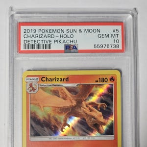 May include: A graded Pok&eacute;mon card featuring Charizard from the 2019 Sun & Moon set. The card is encased in a protective holder with a red label displaying the card's details, including "Detective Pikachu" and a PSA grade of 10.