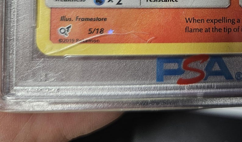 May include: A close-up of a graded trading card encased in a clear plastic holder. The card has a yellow and orange background with text and a logo. The holder has the letters "PSA" in blue and red.