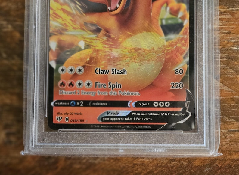 Puede incluir: Una carta coleccionable Pok&eacute;mon Charizard V, en un protector. La carta presenta una ilustraci&oacute;n naranja y amarilla de Charizard. El texto incluye "Claw Slash", "Fire Spin" y "V rule".