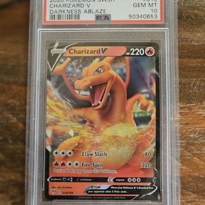 Puede incluir: Una carta Pok&eacute;mon Charizard V graduada del set Darkness Ablaze, dentro de un protector. La carta presenta una ilustraci&oacute;n de Charizard naranja con el texto "Claw Slash" y "Fire Spin". La carta est&aacute; calificada como Gem Mint 10.