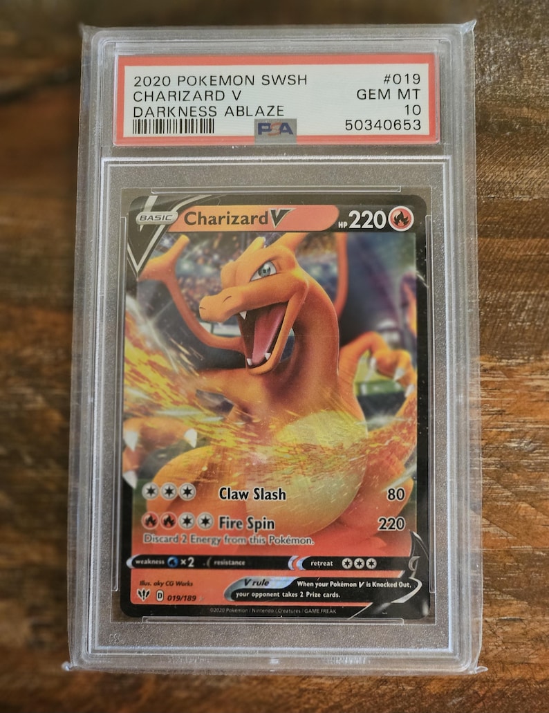 Puede incluir: Una carta Pok&eacute;mon Charizard V graduada del set Darkness Ablaze, dentro de un protector. La carta presenta una ilustraci&oacute;n de Charizard naranja con el texto "Claw Slash" y "Fire Spin". La carta est&aacute; calificada como Gem Mint 10.