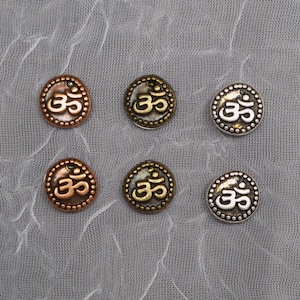 May include: Six metal charms with the Om symbol in different finishes. The charms are silver, gold, and copper.
