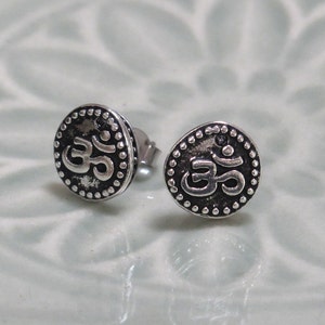 May include: A pair of round silver stud earrings featuring a raised Om symbol in the center. The circular design is bordered by a series of small, raised silver dots, and the background behind the Om symbol has a dark, oxidized finish.