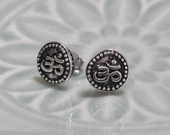 May include: A pair of round silver stud earrings featuring a raised Om symbol in the center. The circular design is bordered by a series of small, raised silver dots, and the background behind the Om symbol has a dark, oxidized finish.