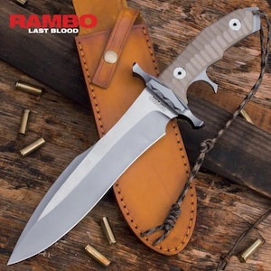 May include: A large, silver-bladed knife with a tan handle and a leather sheath. The knife has the text "RAMBO LAST BLOOD" on the blade. The sheath is brown and has a button closure. Several bullet casings are scattered around the knife.
