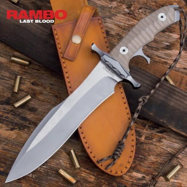 May include: A large, silver-bladed knife with a tan handle and a leather sheath. The knife has the text "RAMBO LAST BLOOD" on the blade. The sheath is brown and has a button closure. Several bullet casings are scattered around the knife.