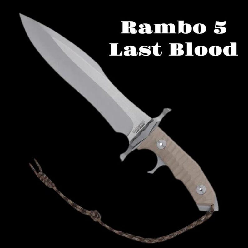 May include: A large, silver knife with a light brown handle and a lanyard. The text "Rambo 5 Last Blood" is displayed above the knife. The knife has a wide blade and a pointed tip.